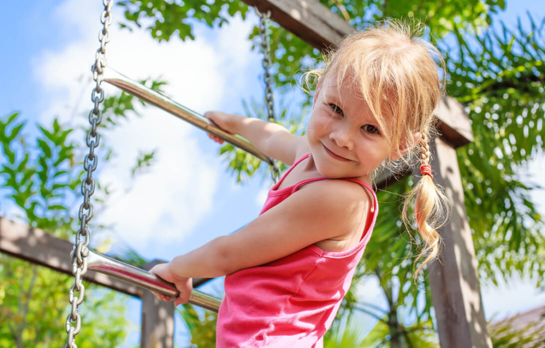 Natural Pharmaceuticals OmegaMarine Junior Mosolygós kislány játszik kültéri mászókán napos időben, háttérben zöld növényekkel és kék égbolt.