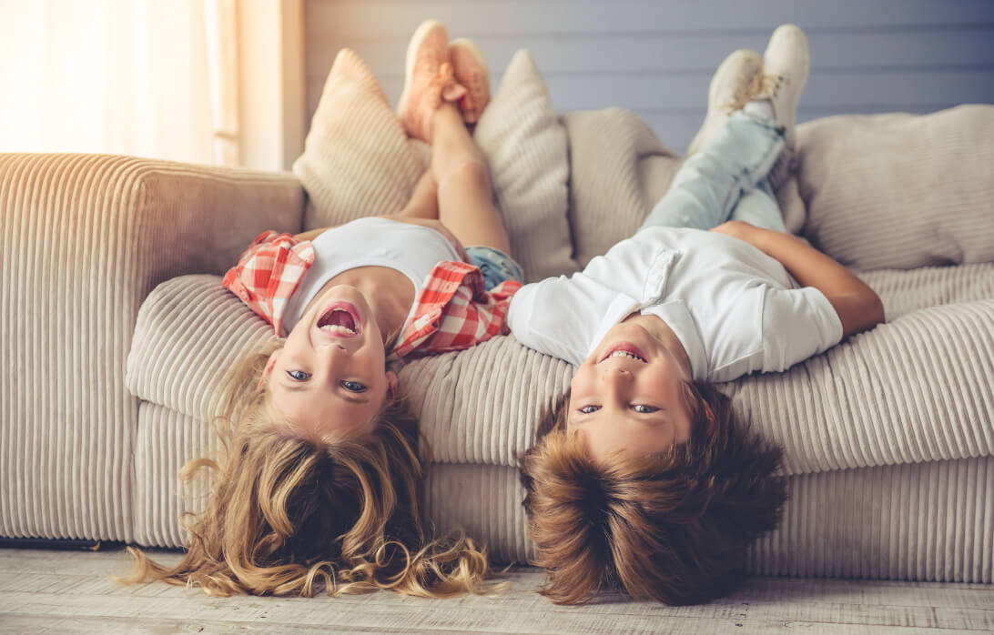 Natural Pharmaceuticals OmegaMarine Junior Két vidám gyerek fejjel lefelé hever a kanapén, természetes fényben, otthoni környezetben.