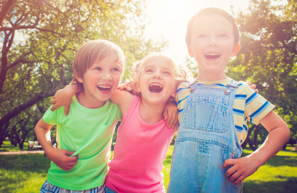 Natural Pharmaceuticals OmegaMarine Junior Három nevető gyermek ölelik egymást egy napfényes parkban, vidám tavaszi hangulatban.