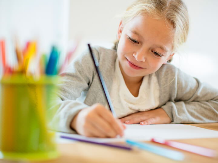 Natural Pharmaceuticals OmegaMarine Junior A kislány színes ceruzákkal rajzol egy fehér papírlapra, arcán öröm és koncentráció látszik.