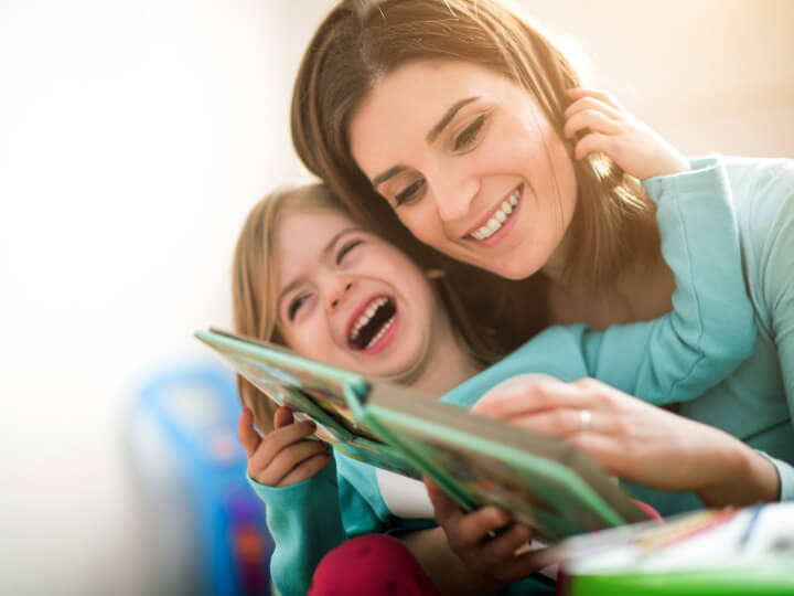 Natural Pharmaceuticals OmegaMarine Junior Boldog anya és kislánya együtt olvasnak egy színes képeskönyvet otthon, örömteli pillanatban.