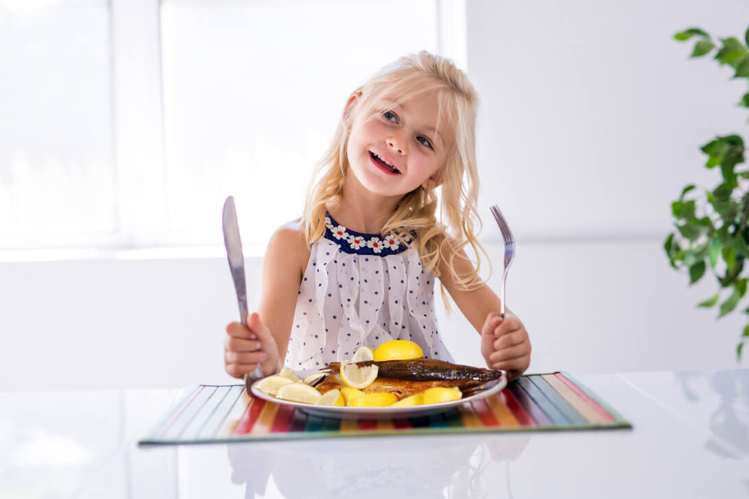 Natural Pharmaceuticals OmegaMarine Junior Vidám kislány kés-villával készen áll a sült hal és citrommal tálalt krumpli fogyasztására egy világos étkezőben.