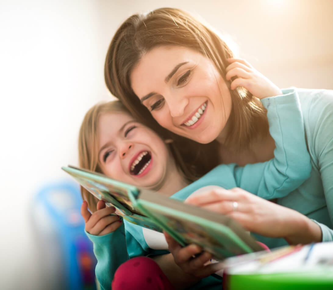 Natural Pharmaceuticals OmegaMarine Junior Mosolygó anya és kislánya együtt olvasnak mesekönyvet otthon, boldog, közös pillanatokat élvezve.