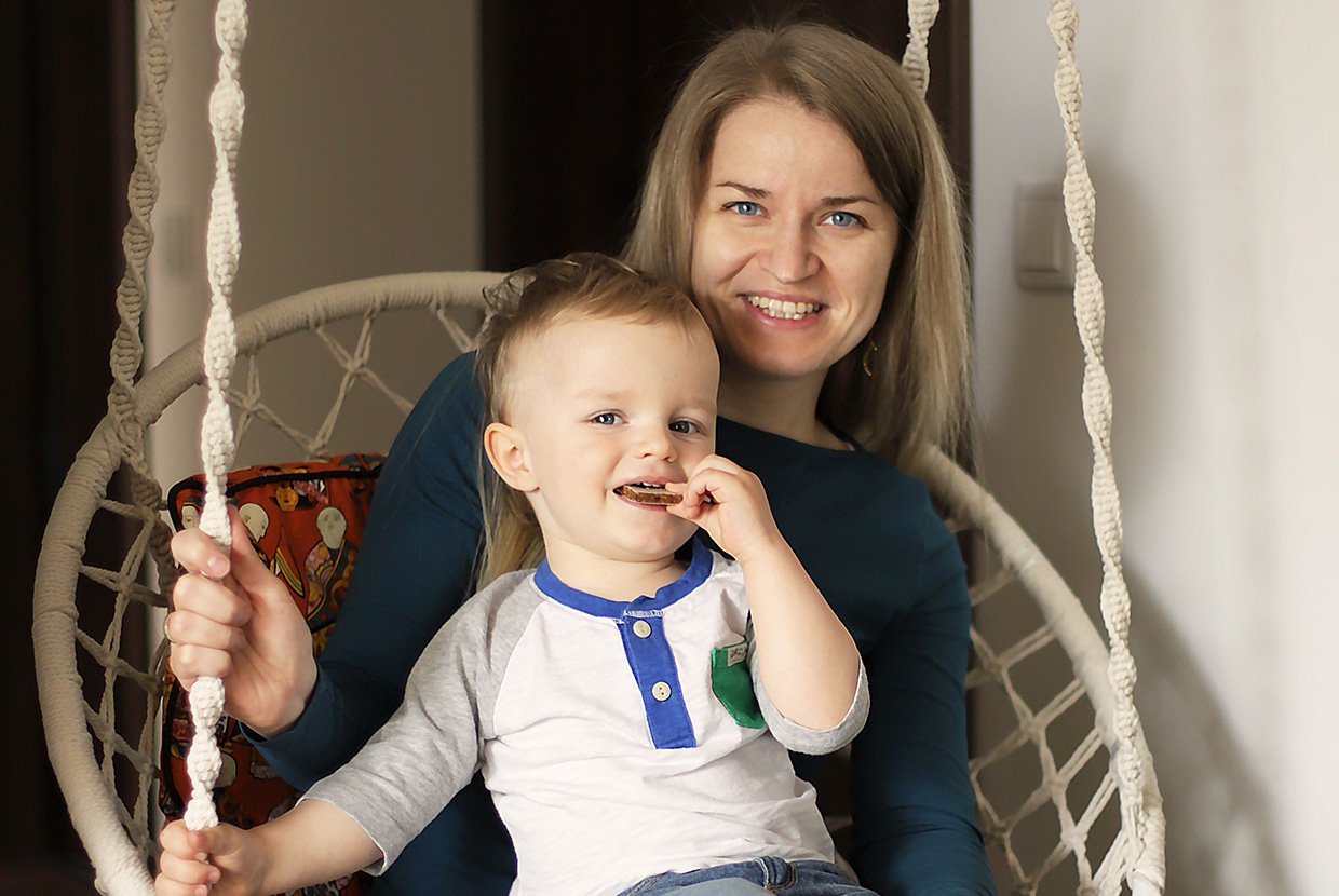 Natural Pharmaceuticals OmegaMarine Junior Mosolygós édesanya egy gyerekkel hintaszékben, a kisfiú sütit majszol.