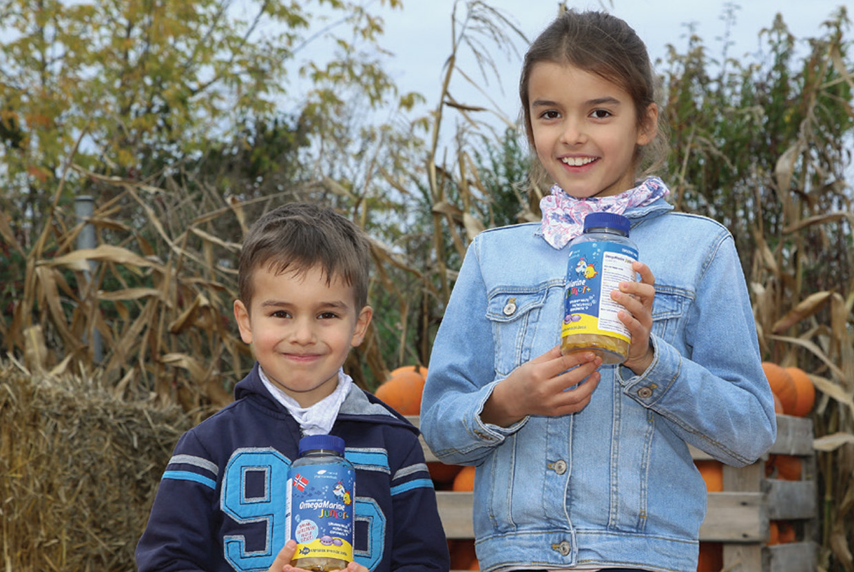 Natural Pharmaceuticals OmegaMarine Junior Két gyermek tart OmegaMarine Junior étrend-kiegészítőt egy őszi tökös kertben.