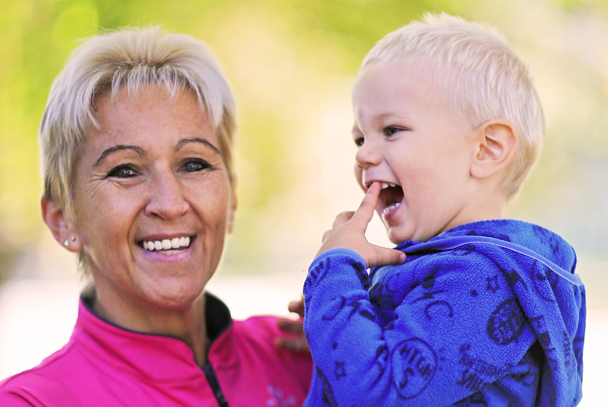 Natural Pharmaceuticals OmegaMarine Junior Boldog nagymama mosolyogva tartja karjában kisunokáját kék pulóverben, zöld háttér előtt.